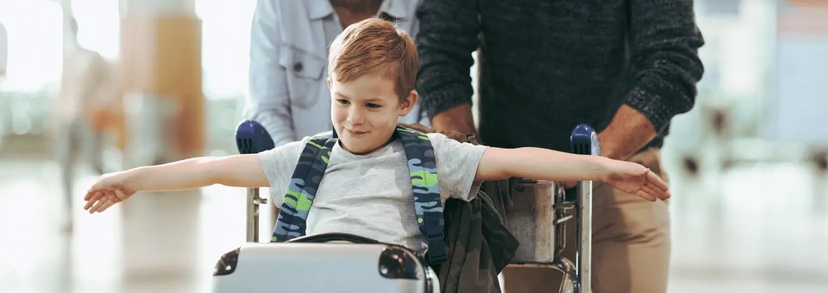 A child is sitting in an airport luggage cart, spreading their arms to mimic an airplane’s wings.